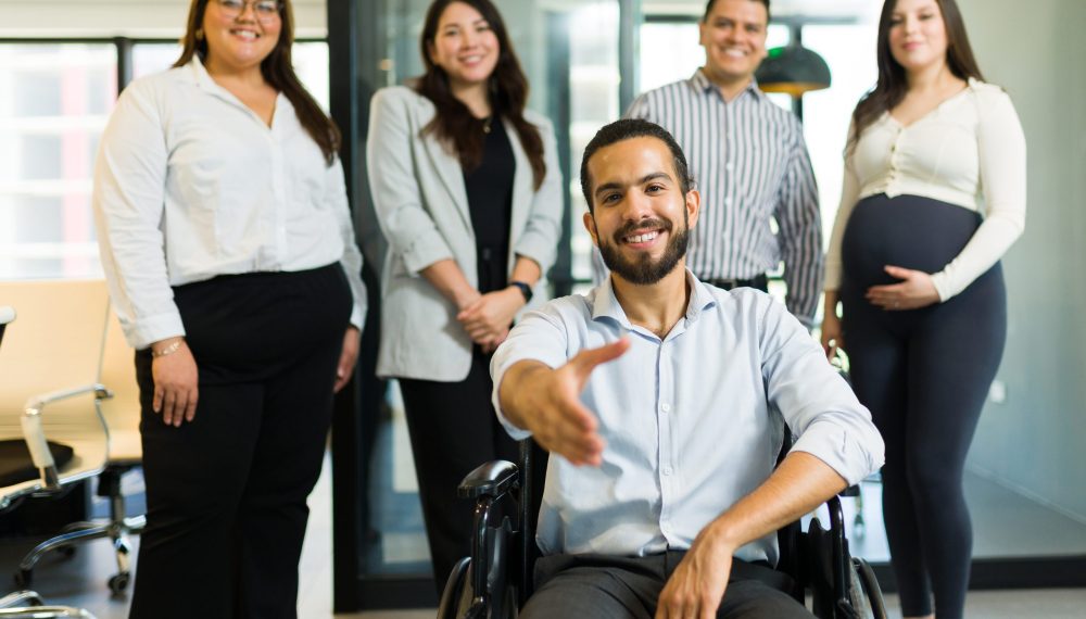 Portrait of a disabled manager sitting on wheelchair offering a handshake with his colleagues standing at back in office