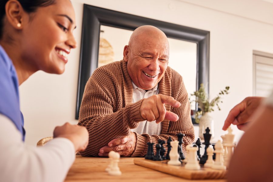 Senior man, nursing home and chess for game, competition and happy with friends, strategy and relax together. Elderly person, nurse and board for contest with mindset, excited smile or point at table